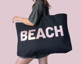 May include: A black canvas tote bag with a pink and white "BEACH" graphic. The bag has a long strap and is being held by a person.