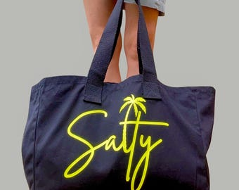 May include: Black tote bag with black handles. The bag features the word "Salty" in a bright yellow script font, with a yellow palm tree graphic above the "y". The bag is held up by a person.