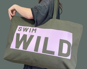 May include: Olive green canvas tote bag with the words "SWIM WILD" printed in white and light purple. The bag has olive green straps and is held by a person.
