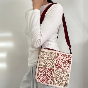 Op de afbeelding: Crèmekleurige crossbodytas met een rood en goudkleurig geborduurd ontwerp. De tas heeft een rechthoekige vorm met een rode band en afwerking. Het geborduurde patroon is een herhalend bloemenontwerp.