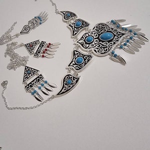Op de afbeelding: Een zilverkleurige sieradenset met een ketting, oorbellen en een armband. De ketting en oorbellen hebben zwart-witte patronen met turkooizen stenen. De armband heeft een vergelijkbaar ontwerp. De set bevat bungelende zilveren en turkooizen accenten.
