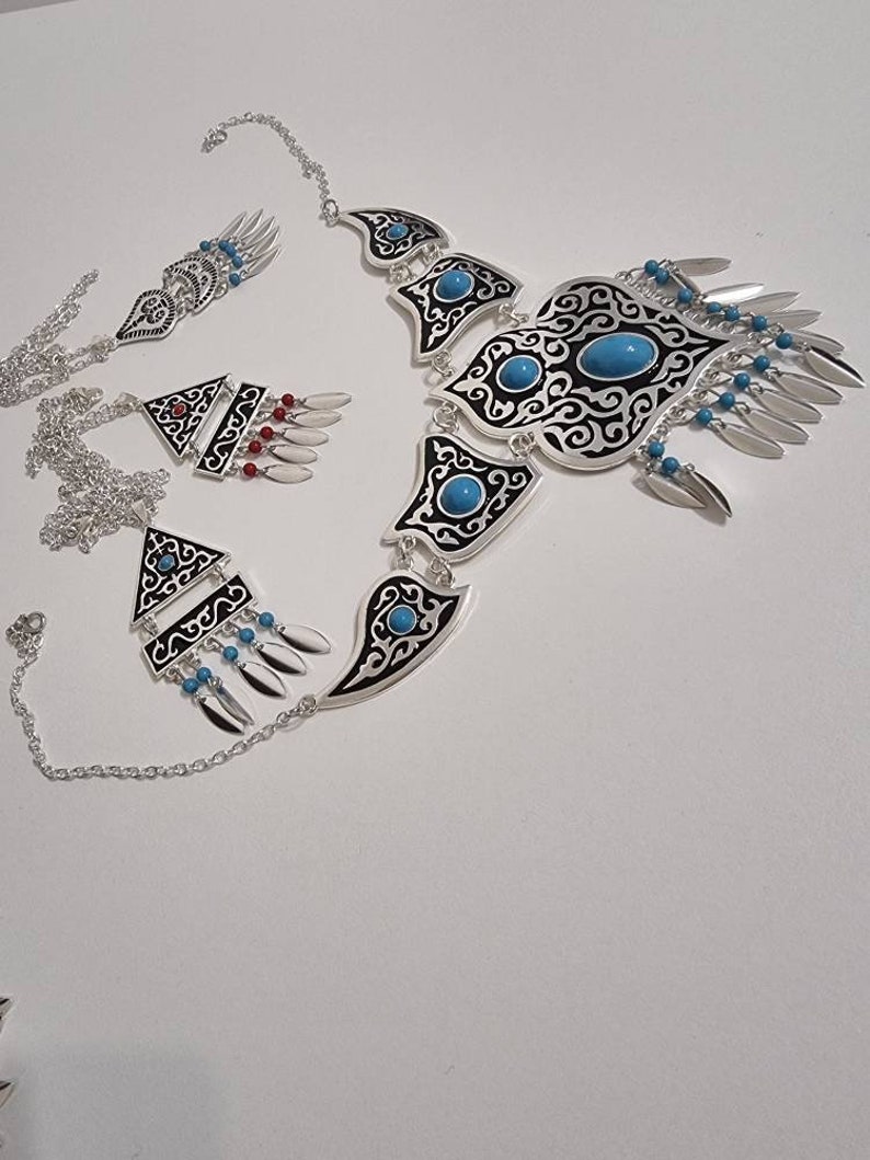 Op de afbeelding: Een zilverkleurige sieradenset met een ketting, oorbellen en een armband. De ketting en oorbellen hebben zwart-witte patronen met turkooizen stenen. De armband heeft een vergelijkbaar ontwerp. De set bevat bungelende zilveren en turkooizen accenten.