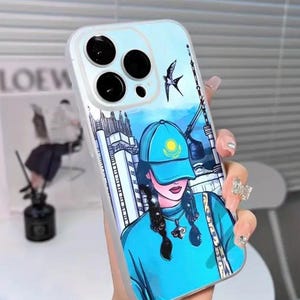 May include: A clear phone case featuring a cartoon illustration of a person wearing a blue cap and sweater, with a cityscape and a bird in the background. The case has a white border and is held by a hand.