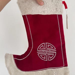 May include: A red Christmas stocking with a silver embroidered design and a fluffy white trim. The stocking has a red ribbon loop and a tag attached. The design is a silver emblem.