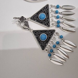 Op de afbeelding: Zilverkleurige oorbellen met een driehoekig zwart en zilver design, met een turquoise middensteen en bungelende zilveren bladvormige bedels. Kleine blauwe kralen accentueren het ontwerp. De oorbellen hebben een decoratieve sluiting.