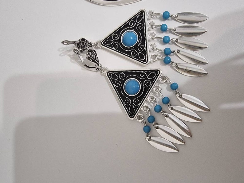 Op de afbeelding: Zilverkleurige oorbellen met een driehoekig zwart en zilver design, met een turquoise middensteen en bungelende zilveren bladvormige bedels. Kleine blauwe kralen accentueren het ontwerp. De oorbellen hebben een decoratieve sluiting.