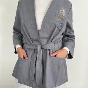 May include: A gray wrap jacket with a V-neck, tie belt, and two front pockets. The jacket features embroidered details on the upper left side. The sleeves are long, and the jacket is worn over a white long-sleeved shirt. The jacket is made of a lightweight fabric.