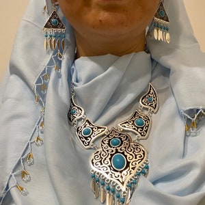 Op de afbeelding: Zilverkleurige sieradenset met een ketting en oorbellen. De ketting heeft een grote hanger met turkooizen stenen en ingewikkelde zwarte en zilveren filigraan. De oorbellen zijn driehoekig met vergelijkbare details. Op een lichtblauwe achtergrond.