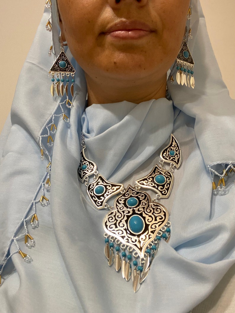 Op de afbeelding: Zilverkleurige sieradenset met een ketting en oorbellen. De ketting heeft een grote hanger met turkooizen stenen en ingewikkelde zwarte en zilveren filigraan. De oorbellen zijn driehoekig met vergelijkbare details. Op een lichtblauwe achtergrond.