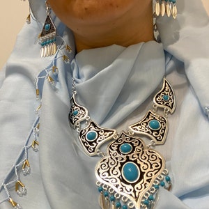 Op de afbeelding: Zilverkleurige ketting en oorbellen set met turquoise accenten. De ketting heeft een sierlijk ontwerp met een grote centrale hanger en bijpassende elementen. De oorbellen zijn driehoekig met bungelende zilveren en turquoise kralen. De sieraden set is tegen een lichtblauwe achtergrond.