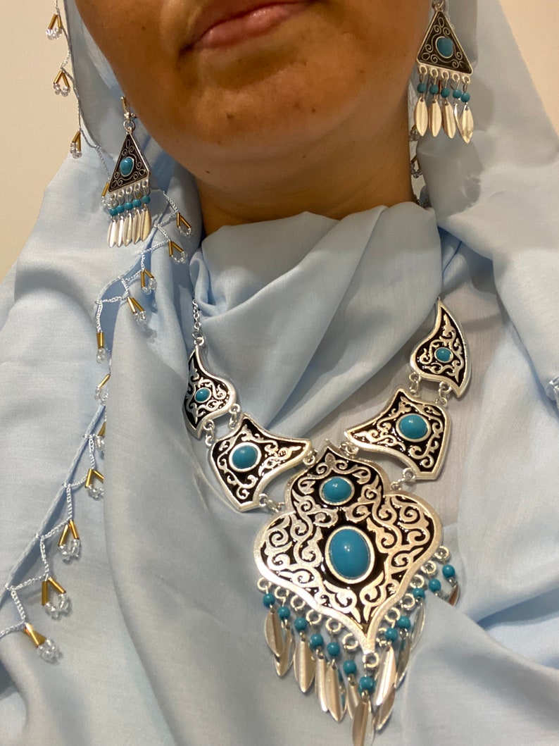 Op de afbeelding: Zilverkleurige ketting en oorbellen set met turquoise accenten. De ketting heeft een sierlijk ontwerp met een grote centrale hanger en bijpassende elementen. De oorbellen zijn driehoekig met bungelende zilveren en turquoise kralen. De sieraden set is tegen een lichtblauwe achtergrond.