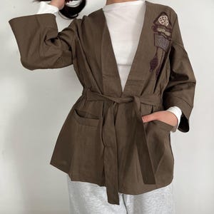 May include: A brown kimono-style jacket with a tie waist and pockets. The jacket features embroidered details on the front and wide sleeves. The sleeves are long and the jacket is open in the front.