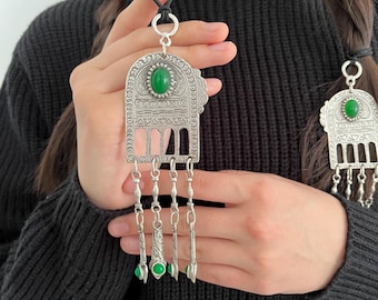 Può includere: Un pendente argentato con una pietra verde ovale e elementi decorativi pendenti. Il pendente ha un design ornato ed è sospeso a un cordino nero. Il pendente è tenuto da una persona.