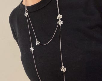 Può includere: Una collana color argento con una catena delicata e molteplici ciondoli decorativi a forma di fiore. La collana è lunga, con un design a strati, e viene indossata su uno sfondo nero.