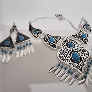 Op de afbeelding: Een zilverkleurige ketting- en oorbellenset. De ketting heeft een grote hanger met zwarte filigraan en turkooizen stenen. De oorbellen zijn driehoekig met vergelijkbare details en bungelende zilveren, bladvormige bedels en turkooizen kralen. De set wordt gepresenteerd op een wit oppervlak.