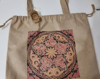 May include: A beige canvas tote bag with a drawstring closure. The bag features a printed design of a star with intricate details in shades of pink, orange, and brown. The text "KAKA" Art Store is printed in blue below the design.
