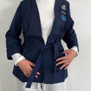 May include: Navy blue kimono-style jacket with a tie belt. The jacket features embroidered patches in shades of blue and beige on the upper left side. It has long sleeves and a relaxed fit. The model is wearing a white long-sleeve shirt underneath.