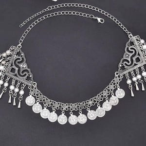Puede incluir: Collar de cadena para el cuerpo plateado con un diseño decorativo. El collar presenta una cadena, elementos triangulares de filigrana, pequeñas cuentas blancas y dijes colgantes en forma de lágrima. También hay pequeños dijes redondos en forma de moneda.