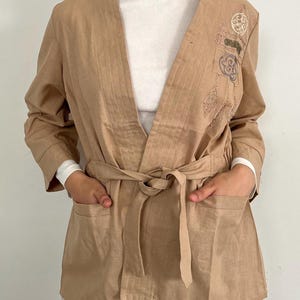 May include: A tan kimono-style jacket with long sleeves, a tie belt, and two front pockets. The jacket features embroidered details on the upper right side. The person is wearing a white long-sleeve shirt underneath.