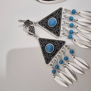 Op de afbeelding: Zilverkleurige oorbellen met een driehoekig zwart-zilveren ontwerp, met een turquoise stenen midden en bungelende zilveren bladvormige bedels. Kleine blauwe kralen accentueren het ontwerp. De oorbellen hebben een decoratieve haak.