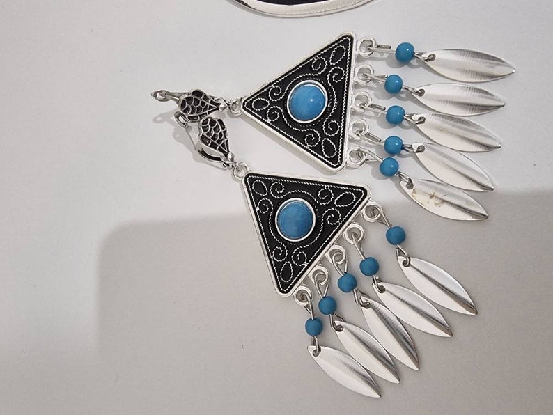 Op de afbeelding: Zilverkleurige oorbellen met een driehoekig zwart-zilveren ontwerp, met een turquoise stenen midden en bungelende zilveren bladvormige bedels. Kleine blauwe kralen accentueren het ontwerp. De oorbellen hebben een decoratieve haak.