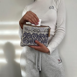 Peut inclure: Une pochette rectangulaire avec une fermeture éclair, ornée d'un motif brodé complexe dans des tons de bleu et de blanc. La pochette est ouverte, révélant un objet blanc à l'intérieur. Une petite étiquette est suspendue à la fermeture éclair.