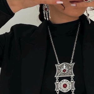 Puede incluir: Juego de joyas plateadas con collar, pendientes y anillos con detalles de piedras preciosas rojas. El collar tiene una cadena larga con un colgante. Los pendientes son colgantes y los anillos tienen un diseño similar. Las joyas se usan con un top y blazer negros.
