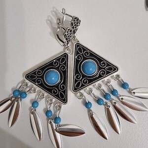 Op de afbeelding: Zilveren oorbellen met een zwarte driehoekige rand en een turquoise middensteen. De oorbellen hebben een decoratief filigraanontwerp en bungelende zilveren, bladvormige bedels met turquoise kralen.