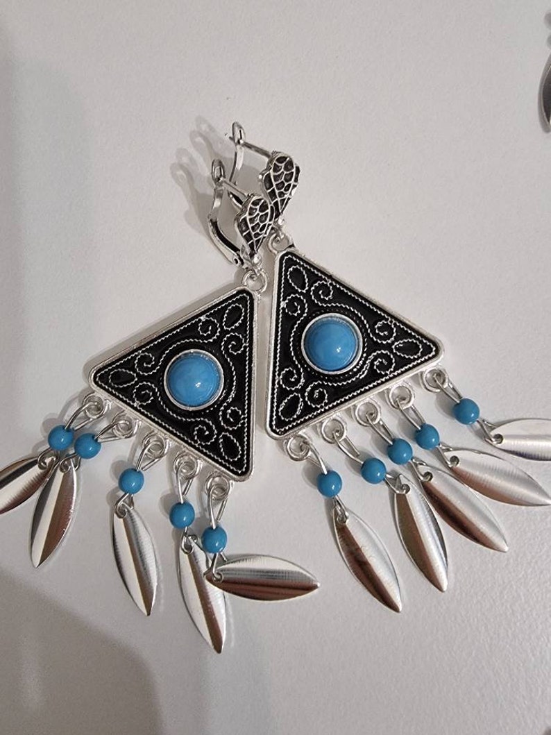 Op de afbeelding: Zilveren oorbellen met een zwarte driehoekige rand en een turquoise middensteen. De oorbellen hebben een decoratief filigraanontwerp en bungelende zilveren, bladvormige bedels met turquoise kralen.