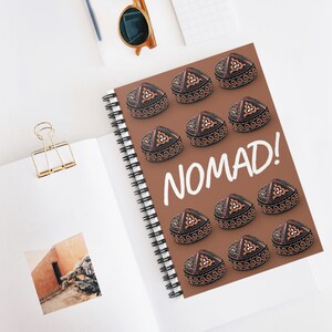 May include: Spiral-bound notebook with a brown cover featuring a pattern of decorative hats and the word "NOMAD!" in white. The hats are black, brown, and orange. The notebook is open on a white surface with a gold paper clip and sunglasses.