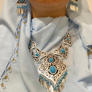 Op de afbeelding: Zilverkleurige ketting en oorbellen set met turquoise stenen en bungelende bladvormige bedels. De ketting heeft een sierlijk ontwerp met zwarte accenten. De driehoekige oorbellen passen bij de ketting. De sieraden worden getoond tegen een lichtblauwe stof.