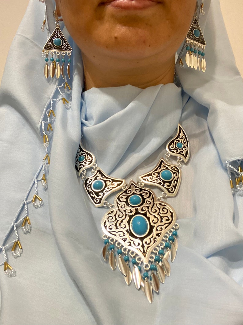 Op de afbeelding: Zilverkleurige ketting en oorbellen set met turquoise stenen en bungelende bladvormige bedels. De ketting heeft een sierlijk ontwerp met zwarte accenten. De driehoekige oorbellen passen bij de ketting. De sieraden worden getoond tegen een lichtblauwe stof.