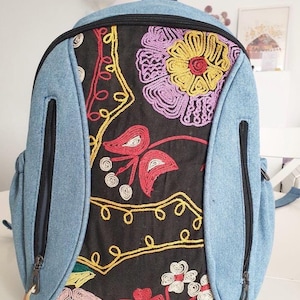 May include: A blue denim backpack with a black fabric panel featuring colorful embroidered flowers. The backpack has two zippered pockets on the front.