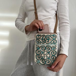 May include: A white and teal embroidered crossbody bag with a brown strap. The bag features an intricate teal and gold floral design. A phone is visible inside the bag. The person is wearing a white long-sleeve shirt and gray sweatpants.