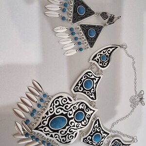 Op de afbeelding: Een zilveren ketting en bijpassende oorbellen set. De ketting heeft zwart-witte filigraan ontwerpen met turkooiskleurige stenen. De oorbellen zijn driehoekig met vergelijkbare details, bungelende zilveren bladvormige bedels en turkoois kralen.