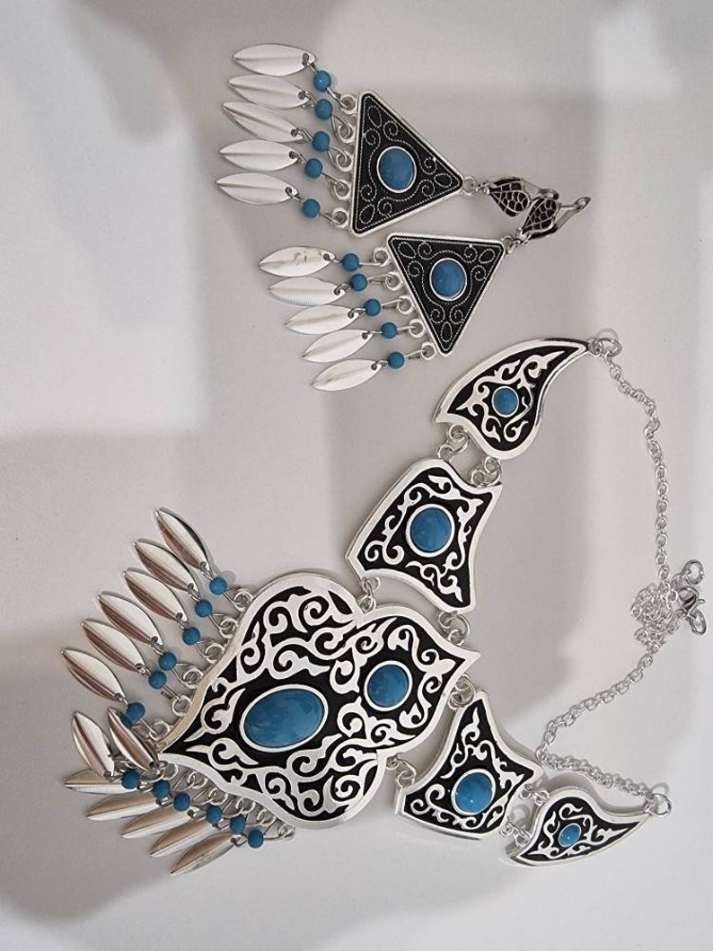Op de afbeelding: Een zilveren ketting en bijpassende oorbellen set. De ketting heeft zwart-witte filigraan ontwerpen met turkooiskleurige stenen. De oorbellen zijn driehoekig met vergelijkbare details, bungelende zilveren bladvormige bedels en turkoois kralen.