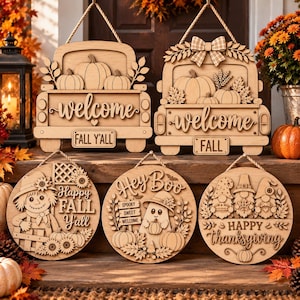 May include: A collection of five wooden hanging signs with laser-cut designs. Two signs are shaped like trucks with pumpkins and the text welcome, FALL Y'ALL, and FALL. Three round signs feature a scarecrow, a ghost with text, and three gnomes.