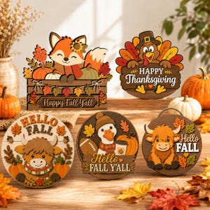 Op de afbeelding: Een set van vijf decoratieve houten bordjes met herfstthema's. De ontwerpen bevatten een vos in een krat met 'Happy Fall Y'all', een kalkoen met 'Happy Thanksgiving', en drie ronde bordjes met 'Hello Fall' of 'Hello Fall Y'all' met een hooglandrund, een gans en een hooglandrund.