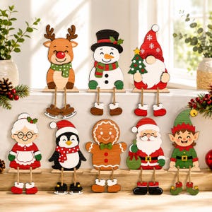 Può includere: Un set di otto decorazioni natalizie in legno da scaffale raffiguranti una renna, un pupazzo di neve, uno gnomo, la signora Claus, un pinguino, un omino di pan di zenzero, Babbo Natale e un elfo, con gambe pendenti realizzate in corda.