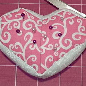 Könnte beinhalten: Ein rosa herzförmiges Nadelkissen mit einem weißen Wirbelmuster und einem weißen Rand. Das Nadelkissen ist mit Füllmaterial gefüllt und hat mehrere Nadeln, die darin stecken.