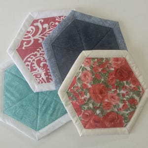 Pode incluir: Quatro bases para copos hexagonais de tecido. Uma apresenta um design paisley vermelho e branco, outra tecido turquesa, uma terceira tecido azul semelhante ao ganga e a última um padrão floral com rosas vermelhas. Cada base tem uma borda branca.