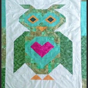 May include: A quilted wall hanging featuring a pieced owl design in shades of green, teal, and white. The owl has orange eyes and a beak, with a bright pink heart centered on its chest, all set against a white background with green borders.