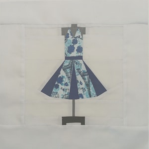 May include: A white dress with a blue and white floral pattern is displayed on a mannequin. The dress has a blue sash around the waist and a blue skirt with a white pattern.