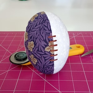 Peut inclure: Un œuf en tissu violet et marron avec une moitié en tissu blanc, cousu ensemble avec du fil marron. L'œuf est posé sur un tapis de coupe rose.