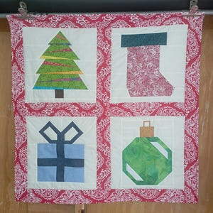 May include: A Christmas-themed quilt with four squares featuring a tree, stocking, gift, and ornament. The tree is green with colorful stripes, the stocking is red with a floral pattern, the gift is blue, and the ornament is green. The quilt has a red and white paisley border.