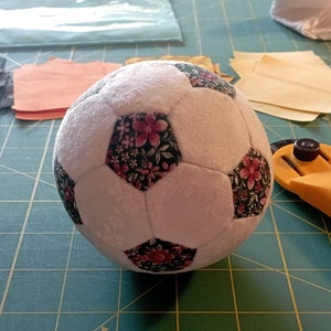 May include: A white football with pink and green floral fabric panels. The ball is sitting on a green cutting mat.