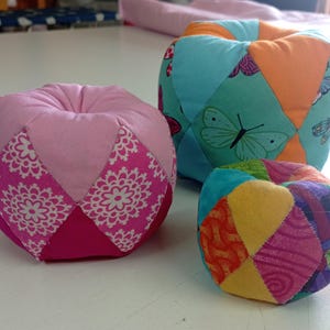 May include: Three colourful fabric balls, each with a different pattern. The largest ball is pink with white floral designs, the middle ball is turquoise and orange with butterfly designs, and the smallest ball is multi-coloured with geometric patterns.