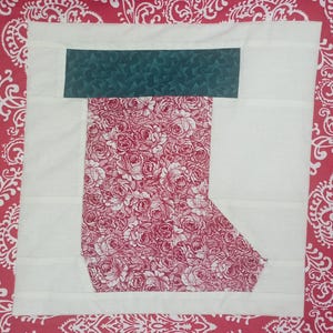 May include: A quilted Christmas stocking block with a white background and a red floral pattern. The top of the stocking is a dark green fabric with a small star pattern. The stocking is a festive holiday craft.
