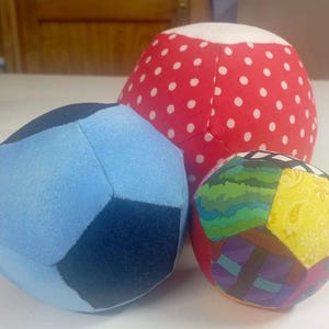 Puede incluir: Tres bolas de tela coloridas. Una es azul claro y azul oscuro, otra es roja con lunares blancos, y la tercera es un patchwork de varios patrones y colores, incluyendo amarillo, verde y morado. Estas bolas blandas son adecuadas para niños.