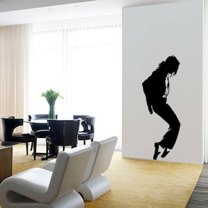 May include: Black and white silhouette wall decal of a person dancing. The decal is applied to a white wall in a living room setting. The room features a white sofa, a dining table with black chairs, and a window with white curtains.